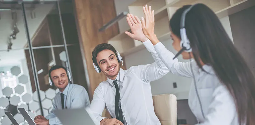 Due operatori con cuffie si danno il cinque in ufficio, con un collega sorridente sullo sfondo