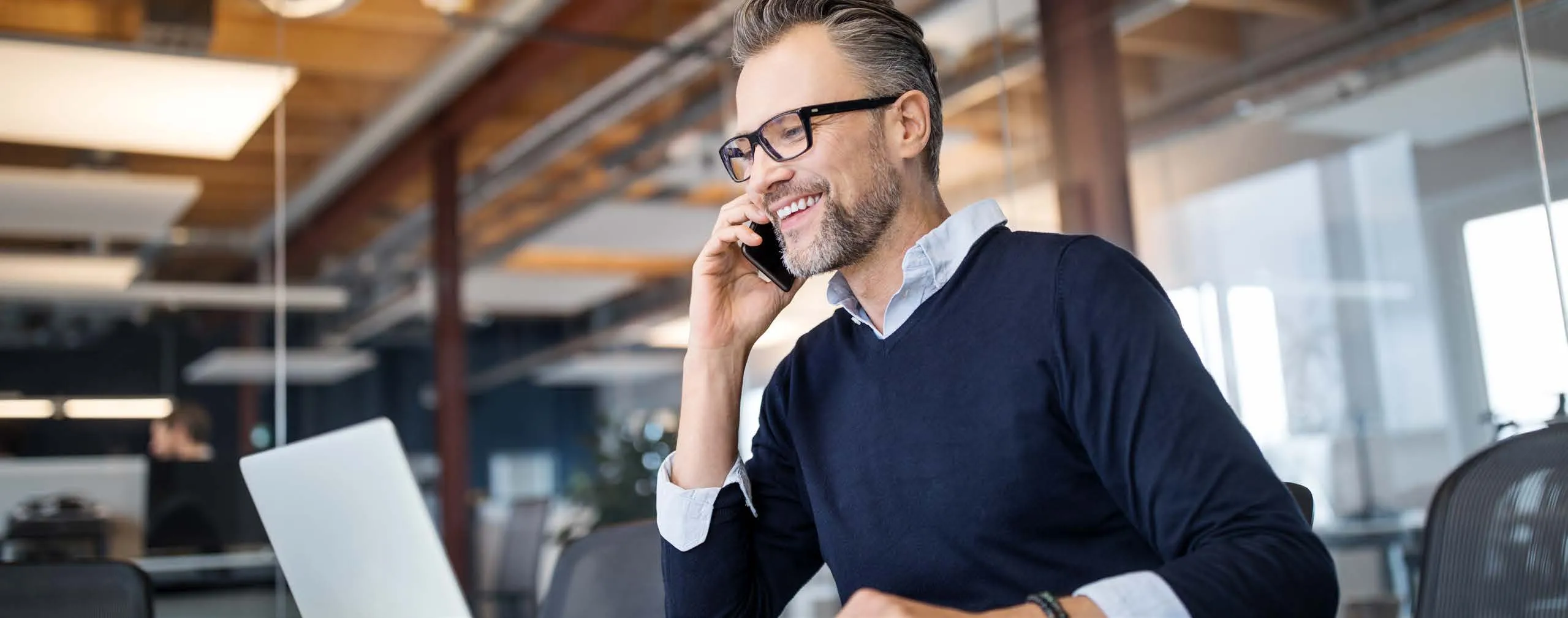 Un uomo con gli occhiali sorride mentre telefona, seduto a una scrivania in un ufficio luminoso con un portatile davanti.