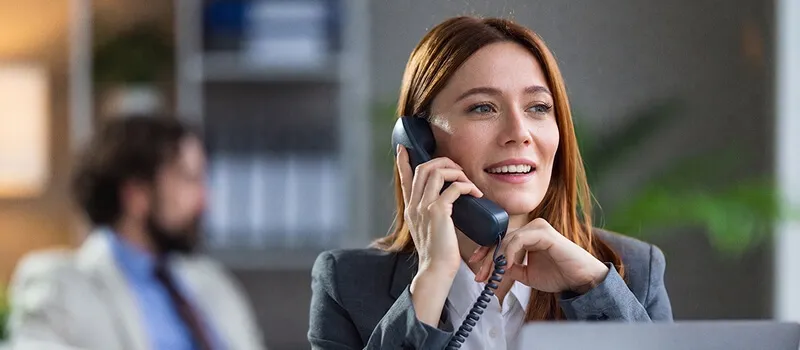 Una donna in giacca grigia risponde sorridendo a una chiamata con un telefono fisso da ufficio, con un collega sfocato sullo sfondo