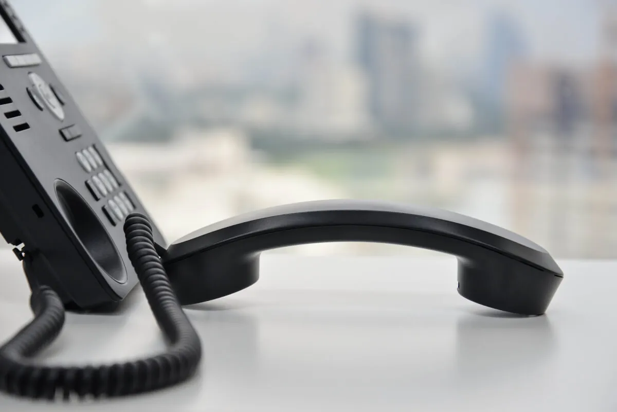 Telefono fisso aziendale nero con la cornetta appoggiata su una scrivania bianca, sullo sfondo una città sfocata